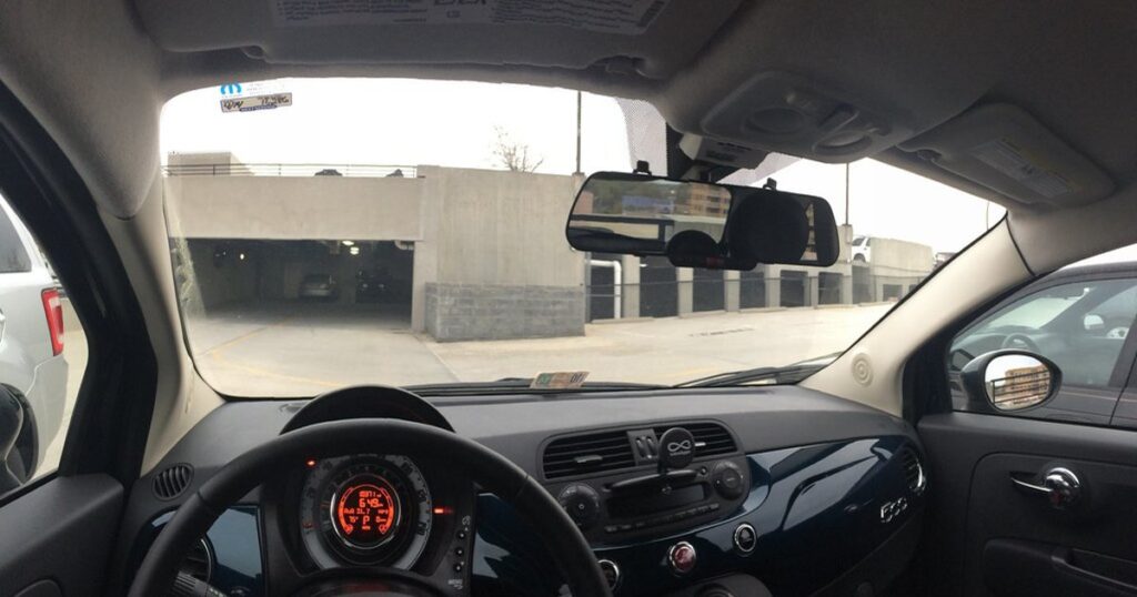 A rearview-mirror-style dash cam mounted across the mirror inside a small car, photographed from the back seat with the windshield and the road ahead visible.